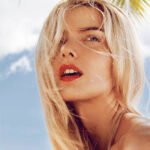 Close-up portrait of beautiful young woman with curly long blonde hair, green mysterious eyes and red lips posing next to the palm.